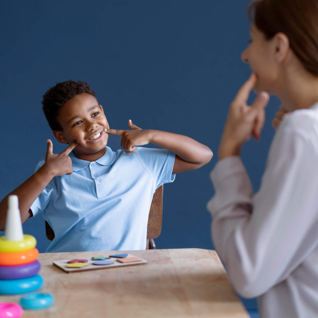 A young boy wearing a blue school uniform, with his fingers on his cheeks as he smiles at the teacher sat opposite him.