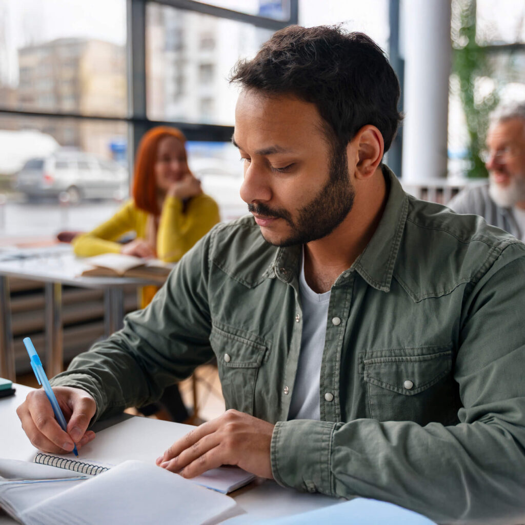 A man wearing a army green jacket sat at a desk, focused on writing in his notebook