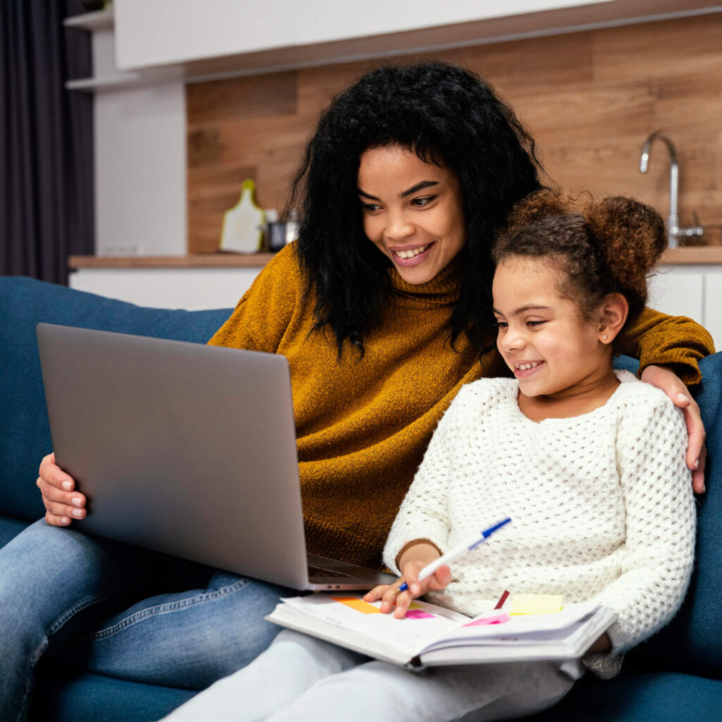 A young mother, wearing a yellow jumper, sat on the sofa with her young daughter. They are smiling, taking notes and looking at their laptop