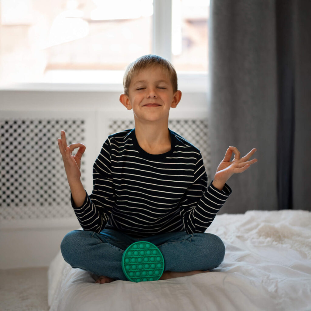 A young boy sat on his bed, wearing a striped jumper, meditating and smiling with a green fidget toy rested against his legs