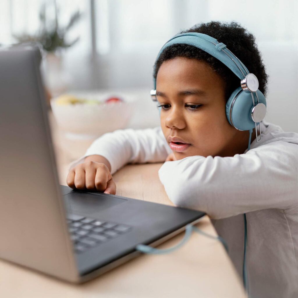 A young boy, sat at his desk wearing blue headphones, focused and relaxed while he looks at his laptop screen