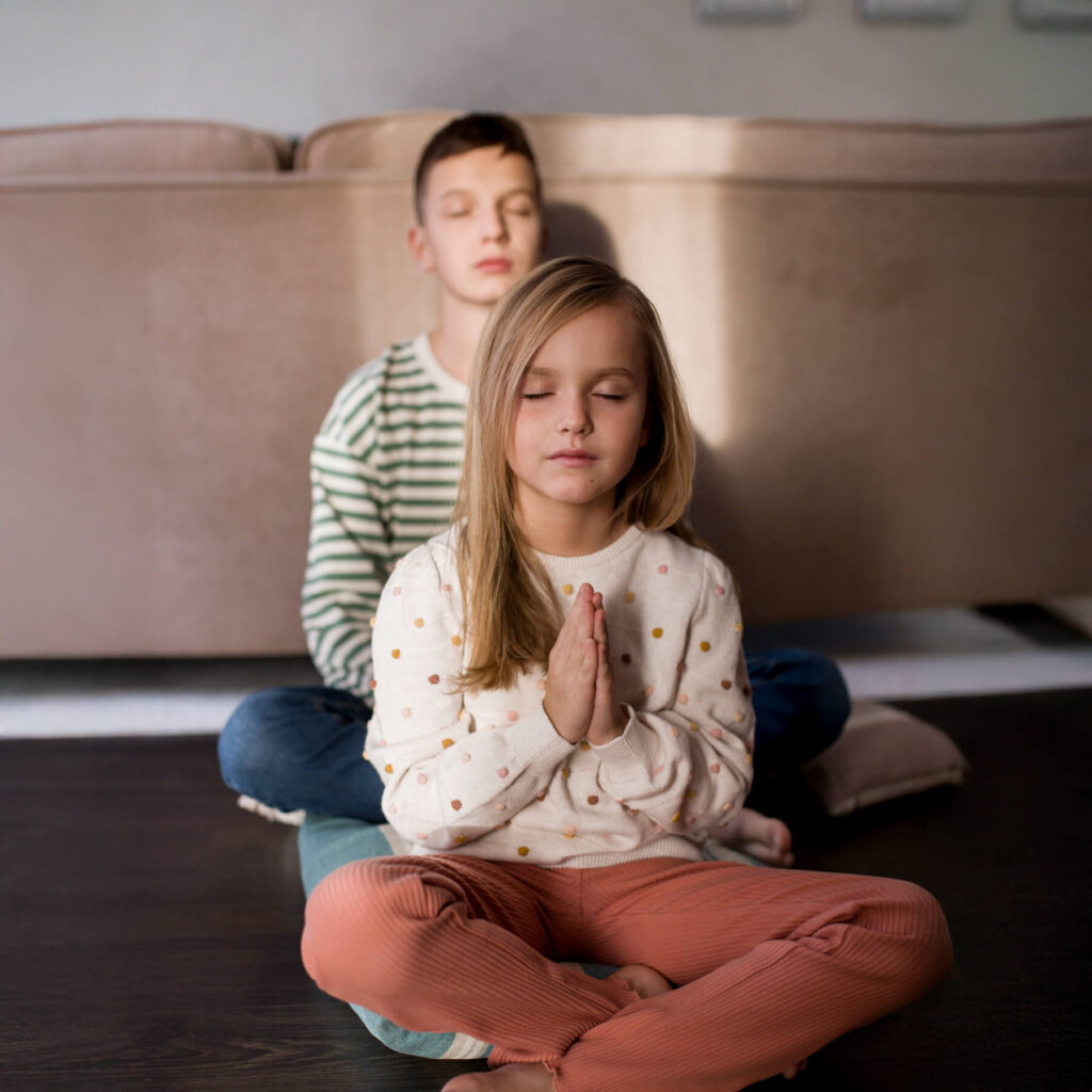 Two children sat on the floor, leaning against a sofa, with their eyes closed meditating