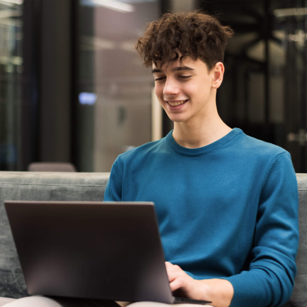 A young man wearing a blue jumper, sat on the sofa smiling while looking at his laptop.