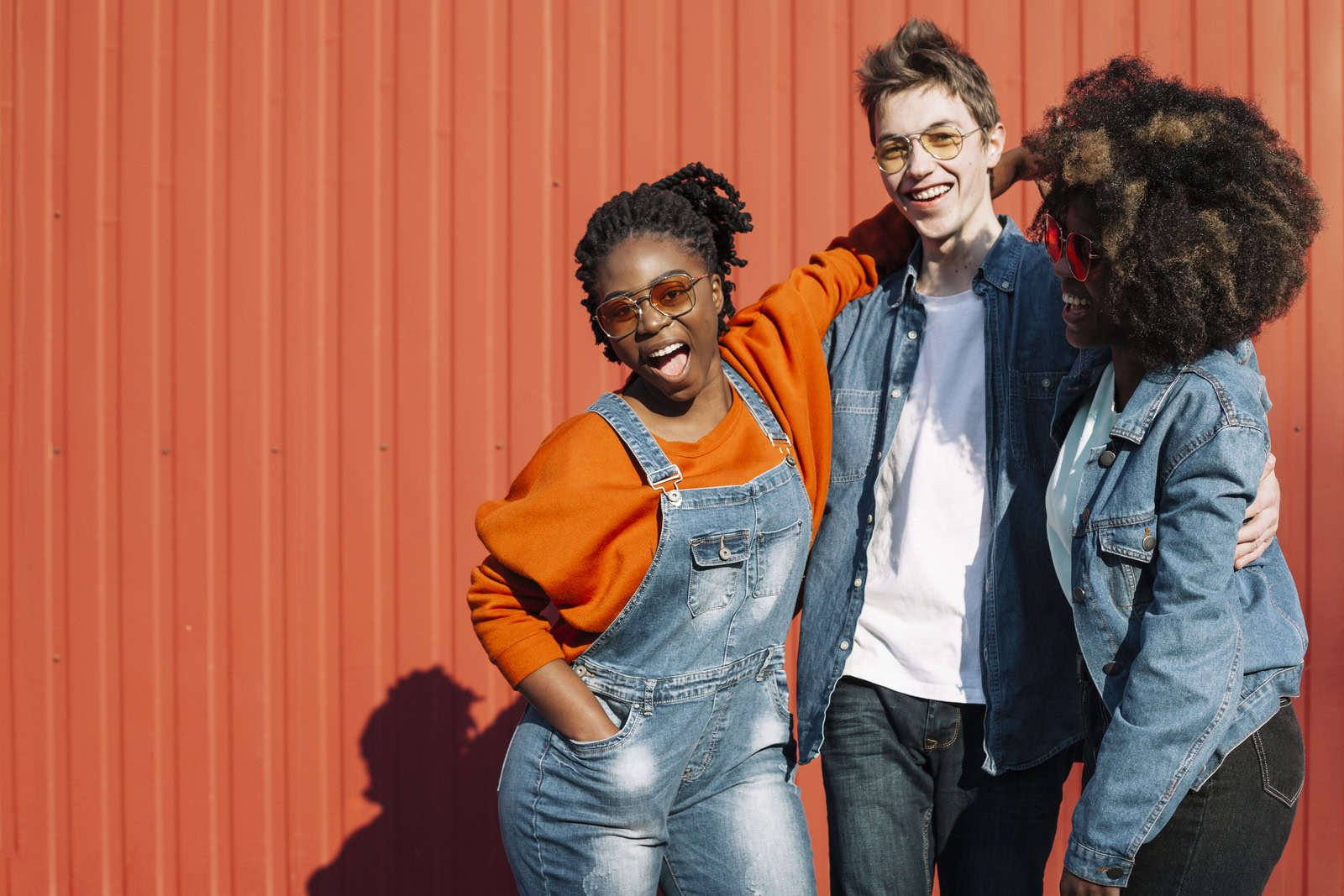 Three young adults are stood outside, by an orange wall, all laughing and enjoying themselves